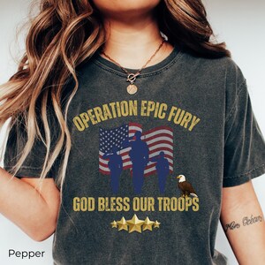 May include: Dark gray t-shirt with the text "OPERATION EPIC FURY" above an American flag and silhouettes of soldiers. Below, it reads "GOD BLESS OUR TROOPS" with gold stars and an eagle.