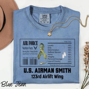 May include: A light blue Comfort Colors t-shirt with the text "AIR FORCE" and "U.S. AIRMAN SMITH". The shirt features a nutrition facts label with military-themed graphics and text. A gold star pendant necklace is draped on the shirt.