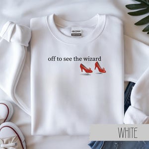 May include: White sweatshirt with the text "off to see the wizard" and an illustration of red shoes. The sweatshirt has a crew neck and long sleeves. A pair of white sneakers are visible in the lower left corner.
