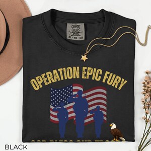 May include: Black t-shirt with the text "OPERATION EPIC FURY" in gold lettering. The design includes an American flag, saluting silhouettes, and an eagle. A gold star pendant necklace rests on the shirt.