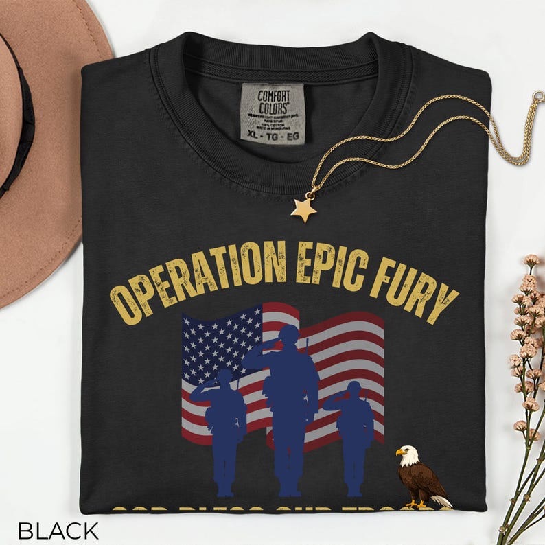 May include: Black t-shirt with the text "OPERATION EPIC FURY" in gold lettering. The design includes an American flag, saluting silhouettes, and an eagle. A gold star pendant necklace rests on the shirt.