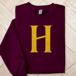 Könnte beinhalten: Burgunderfarbenes Sweatshirt mit einem großen, goldfarbenen, gestrickten Buchstaben "H" auf der Vorderseite. Das Sweatshirt ist gefaltet, wobei der Ärmel sichtbar ist. Das Wort "MAROON" befindet sich in der oberen linken Ecke.