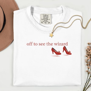 May include: White Comfort Colors t-shirt with the text "off to see the wizard" in red, along with a pair of red high-heeled shoes. A gold necklace with a star pendant and a brown hat are also visible.