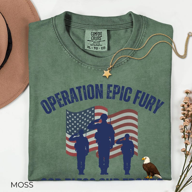 May include: Moss green t-shirt with the text "OPERATION EPIC FURY" above an American flag and silhouettes of soldiers saluting. A gold star pendant necklace and a brown hat are also visible.
