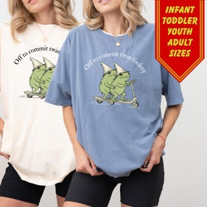 May include: Two t-shirts, one cream and one blue, each featuring a whimsical illustration of two green frogs wearing hats and riding a scooter. The text "Off to commit twin foolery" is arched above the frogs. A red banner indicates sizes.