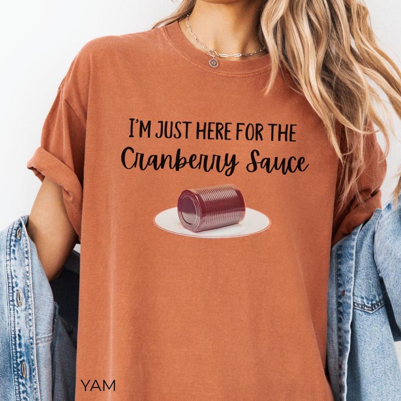 Puede incluir: Camiseta color &oacute;xido con el texto negro "I'M JUST HERE FOR THE Cranberry Sauce". Se muestra una lata de salsa de ar&aacute;ndanos en un plato blanco. La camiseta tiene las mangas dobladas.