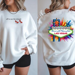 May include: White sweatshirt with the text "off to see the wizard" and a graphic of red high-heeled shoes. The back of the sweatshirt features a colorful "Welcome to Las Vegas" sign design with the phrase "There's No Place Like Las Vegas."