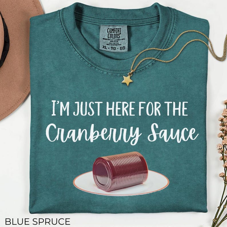 Puede incluir: Camiseta azul abeto con el texto "I'M JUST HERE FOR THE Cranberry Sauce". La camiseta presenta una lata de salsa de ar&aacute;ndanos en un plato. Tambi&eacute;n se ve un collar dorado con un colgante de estrella.
