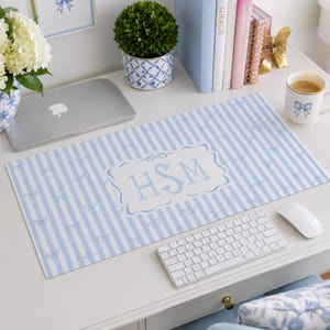 May include: A rectangular desk mat featuring a light blue and white vertical striped pattern with small blue bows scattered throughout. The center displays a white decorative frame containing the blue monogram letters HSM in a serif font.