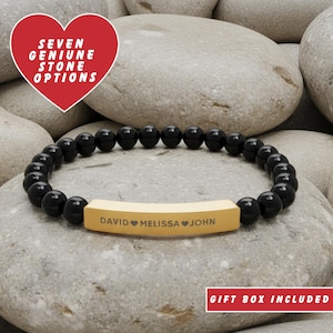 May include: A black beaded bracelet with a gold-coloured bar engraved with names and hearts. The bracelet is displayed on a bed of grey stones. Text in the image reads "SEVEN GENUINE STONE OPTIONS" and "GIFT BOX INCLUDED".