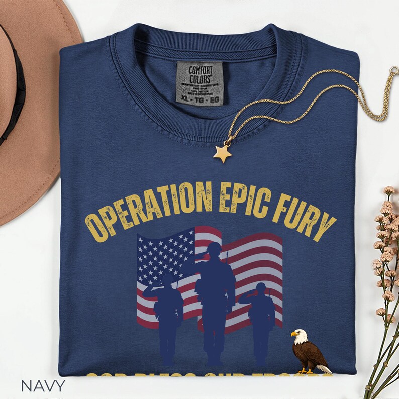 May include: Navy blue t-shirt with the text "OPERATION EPIC FURY" in yellow, featuring an American flag and silhouettes of soldiers saluting. A gold star pendant necklace and a brown hat are also visible.