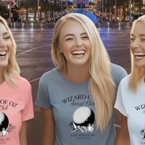 May include: Three women wearing t-shirts with the text "WIZARD OF OZ Social Club" and a golf ball graphic. The shirts are in pink, gray, and white. The text "LAS VEGAS, NV" is printed below the graphic.