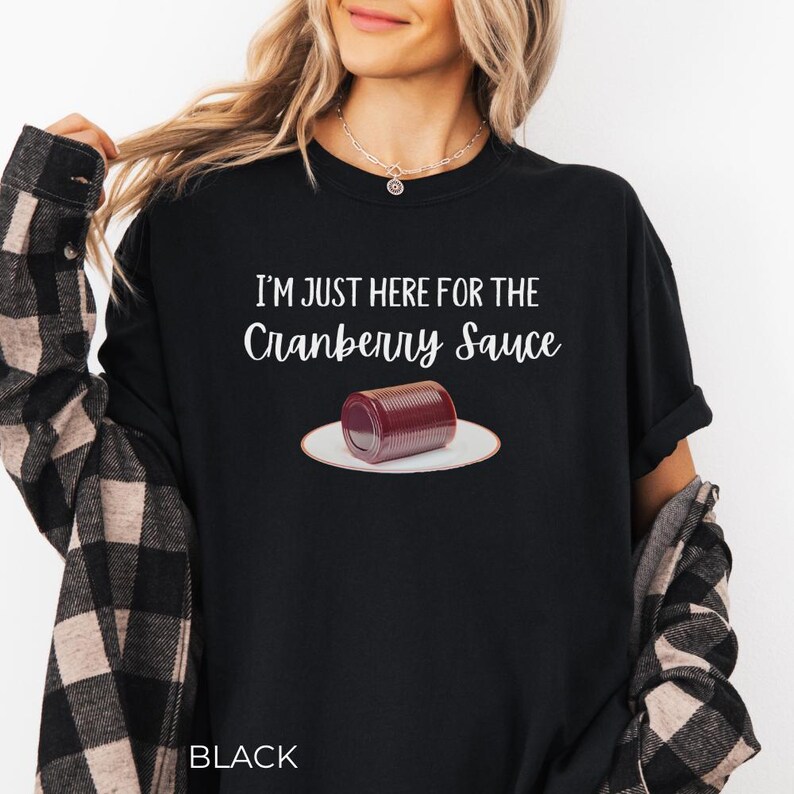 Puede incluir: Camiseta negra con el texto blanco "I'M JUST HERE FOR THE Cranberry Sauce". Un gr&aacute;fico de una lata de salsa de ar&aacute;ndanos en un plato blanco est&aacute; debajo del texto. La palabra "BLACK" est&aacute; en la parte inferior de la camiseta.