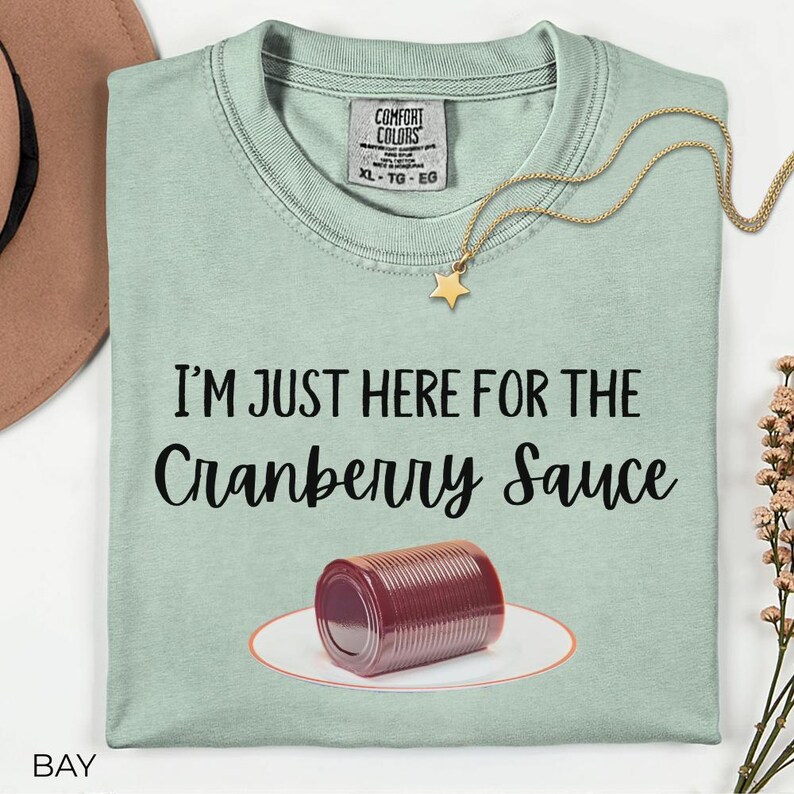 Puede incluir: Camiseta verde claro con el texto "I'M JUST HERE FOR THE Cranberry Sauce" y un gr&aacute;fico de una lata de salsa de ar&aacute;ndanos en un plato. Un collar dorado con un colgante de estrella est&aacute; sobre la camiseta.