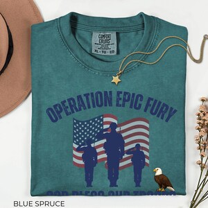 May include: Blue spruce t-shirt with the text "OPERATION EPIC FURY" and an illustration of soldiers saluting in front of an American flag. A gold star necklace and a brown hat are also visible.