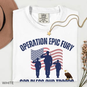 May include: White t-shirt with the text "OPERATION EPIC FURY" above an American flag graphic and saluting soldiers. Below the flag is the text "GOD BLESS OUR TROOPS" and an eagle. A gold star necklace is draped on the shirt.