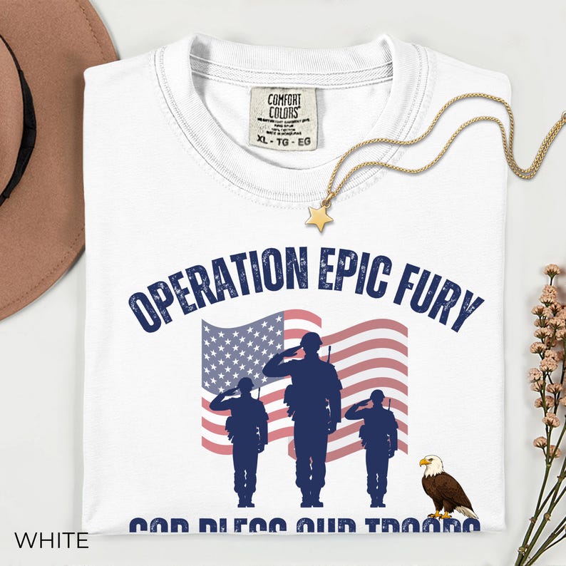 May include: White t-shirt with the text "OPERATION EPIC FURY" above an American flag graphic and saluting soldiers. Below the flag is the text "GOD BLESS OUR TROOPS" and an eagle. A gold star necklace is draped on the shirt.
