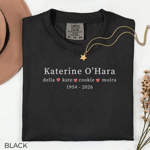 May include: Black crew neck t-shirt with the name "Katerine O'Hara" in white, along with names and dates. A gold star pendant necklace rests on the shirt. The shirt has a Comfort Colors tag and the word "BLACK" at the bottom.