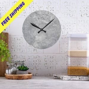 Könnte beinhalten: Eine runde Wanduhr mit grauem Marmor-Effekt-Zifferblatt und schwarzen Zeigern. Die Uhr ist an einer weißen Fliesenwand befestigt. Das Bild zeigt auch ein gelbes Banner mit der Aufschrift "FREE SHIPPING".
