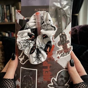 Puede incluir: Un set de regalo de terror que incluye una goma para el pelo blanca con estampado de telaraña negra, un brillo de labios transparente y un cuchillo de plástico en una funda. El paquete tiene salpicaduras de sangre, una máscara de hockey y el texto "FORGIVE KILLER".