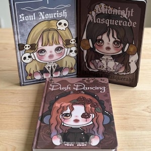 May include: Three hardcover journals featuring illustrated characters on the covers. The left journal is titled Soul Nourish with a blonde character and skulls. The right journal is titled Midnight Masquerade. The front journal is titled Dusk Dancing.