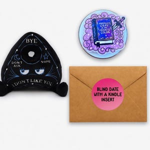 May include: An assortment of novelty items, including a black Ouija board plush with the text "I DON'T LIKE YOU", a round sticker with a book and sword, and a brown envelope with a pink sticker that reads "BLIND DATE WITH A KINDLE INSERT."