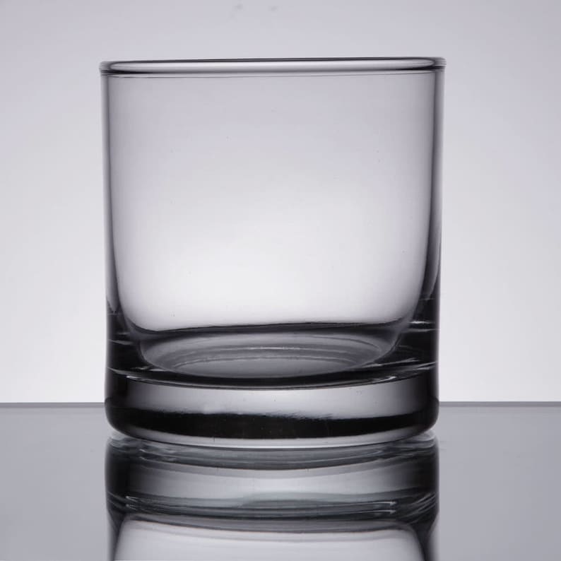 May include: A clear glass tumbler with a thick base. The glass is empty and has a slight reflection of the background.