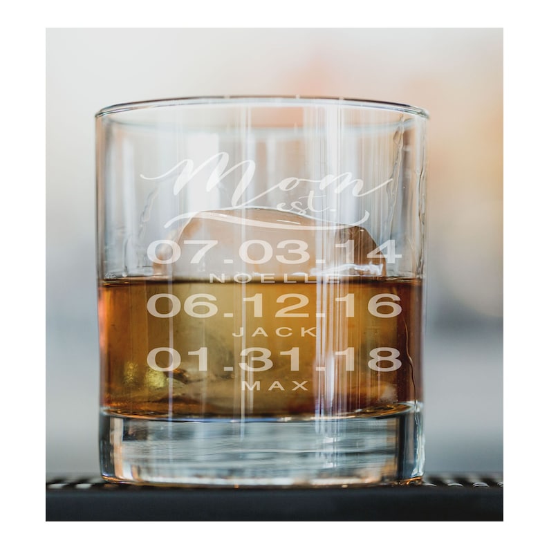 May include: A clear glass with a brown liquid and ice inside. The glass is engraved with the word "Mom" and dates: 07-03-14, 06-12-16, 01-31-18.