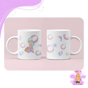 Puede incluir: Dos tazas de cerámica blanca con diseños de sirenas y océanos. Una taza presenta una sirena, burbujas y coral. La otra taza tiene burbujas, un delfín y una estrella de mar. Las tazas tienen un fondo rosa claro.