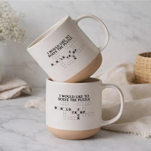 May include: Two white ceramic mugs with a tan base are stacked. Each mug has a black crossword puzzle design and the text "I WOULD LIKE TO SOLVE THE PUZZLE." The mugs have large, curved handles.