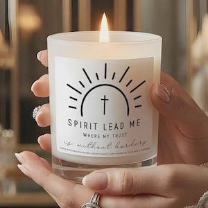 Puede incluir: Una vela blanca encendida en un tarro de cristal transparente. La vela tiene una etiqueta con el texto "SPIRIT LEAD ME" y "WHERE MY TRUST is without borders". La etiqueta también presenta un diseño de sol con una cruz en el centro.