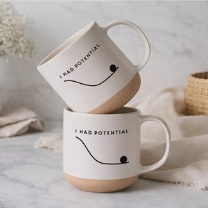 May include: A white ceramic mug with a black handle and the text "But First, Coffee" in a bold, black font. The mug is set against a blurred background, suggesting a cosy setting.