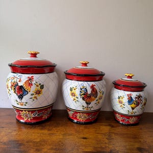 May include: Set of three ceramic kitchen canisters with lids. The canisters are white with a blue diamond pattern, and feature rooster and floral designs. The lids are red with a black trim and yellow finials. The canisters are on a wooden surface.