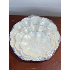 May include: A set of three nested, scalloped-edge plates with a pearlescent, off-white finish. The plates are stacked in descending order of size and are encased in a clear, crinkled plastic wrapping that covers the entire surface of the items.