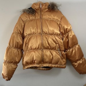 Op de afbeelding: Een gouden pufferjack met een met bont afgezette capuchon. De jas heeft een ritssluiting aan de voorkant en een gewatteerd ontwerp. De mouwen zijn lang en de jas is gemaakt van een glanzend materiaal.