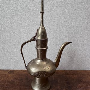 Puede incluir: Una jarra de metal en tono latón con una base bulbosa, un cuello delgado y un pico largo y curvado. La superficie está decorada con patrones grabados de hojas repetidas. Incluye un asa fina y arqueada y una tapa alta y puntiaguda.