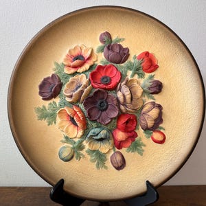 May include: A decorative ceramic plate featuring a raised floral design. The flowers are in shades of red, purple, and yellow, with green leaves. The plate has a tan background and a brown rim. The plate is displayed on a black stand.