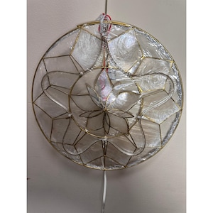 May include: A circular wall hanging featuring a gold-toned metal frame with a geometric star pattern. The structure is filled with translucent, iridescent white shell panels. The entire piece is encased in a clear plastic protective film.