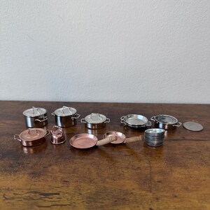 Op de afbeelding: Een verzameling miniatuur metalen kookgerei, waaronder potten, pannen en deksels. De set bevat zowel zilver- als koperkleurige stukken, met kleine handvatten en een stapel miniatuur borden. De items zijn gerangschikt op een houten oppervlak.