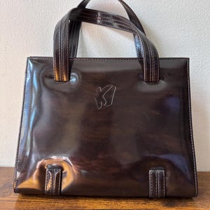 May include: A dark brown, glossy patent leather handbag with a structured, rectangular silhouette. The bag features two top handles, stitched detailing, and the letters KI embossed in the centre of the front panel. The finish is highly reflective.