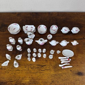 May include: A collection of miniature porcelain tea party dishes. The set includes teacups, saucers, teapots, creamers, sugar bowls, and serving dishes. Each piece is white with a delicate pink floral pattern and gold trim.