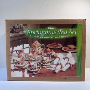 Puede incluir: Un juego de té de 19 piezas Springtime Tea Set en una caja de cartón con acabado de cerámica pintada a mano. La caja muestra una tetera, tazas, platillos y servilleteros con motivos florales y animales, junto a platos con galletas.