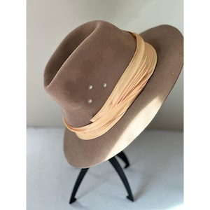 May include: A taupe felt fedora hat featuring a wide, pleated peach-coloured satin ribbon band around the crown. The hat includes three small, circular silver-toned metal ventilation eyelets on the side of the crown, positioned above the ribbon.