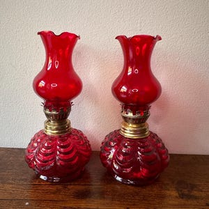 May include: A pair of antique red glass oil lamps. Each lamp has a textured, round base, a brass collar, and a flared top. The lamps are displayed on a wooden surface.