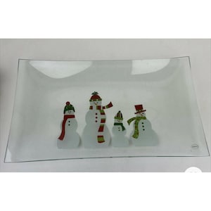 May include: A rectangular clear glass serving platter featuring a festive graphic of four snowmen. Each snowman wears a colourful hat and scarf in shades of red, green, and white. The glass has a smooth, slightly curved edge design and a small China label.
