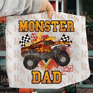 May include: White tote bag featuring a monster truck design with the words "MONSTER DAD" in orange and yellow. The truck has a red and yellow color scheme with checkered flags and a dollar sign.