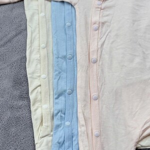 May include: A close-up view of four baby onesies in pastel colors: cream, light yellow, light blue, and pale pink. Each onesie has a row of white snap closures down the front. The fabric appears soft and lightweight, suitable for infants.