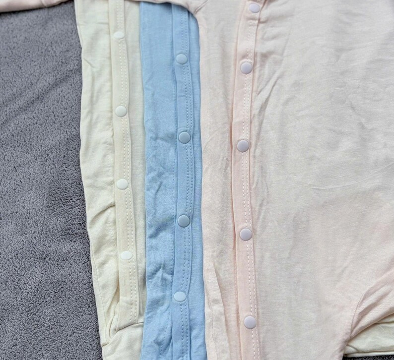 May include: A close-up view of four baby onesies in pastel colors: cream, light yellow, light blue, and pale pink. Each onesie has a row of white snap closures down the front. The fabric appears soft and lightweight, suitable for infants.