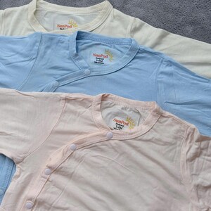 May include: Three pastel-colored baby bodysuits. The bodysuits are light yellow, light blue, and light pink. Each has a wrap-style front with snap closures and a round neckline. The brand name "SunPod" is printed on the neckline.