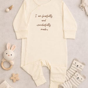 May include: A cream-coloured baby sleepsuit with long sleeves and popper fastenings. The text "I am fearfully and wonderfully made" is printed on the front. A crocheted bunny rattle, star-shaped wooden toy, and striped bear socks are also visible.